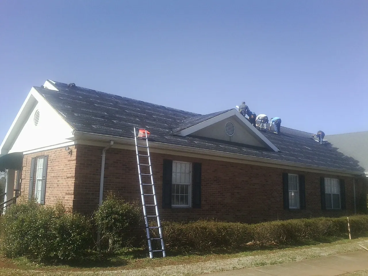Crew tearing shingles off a brick ranch roof with a ladder set — Emergency Roof Repair work by our crew in Romayor
