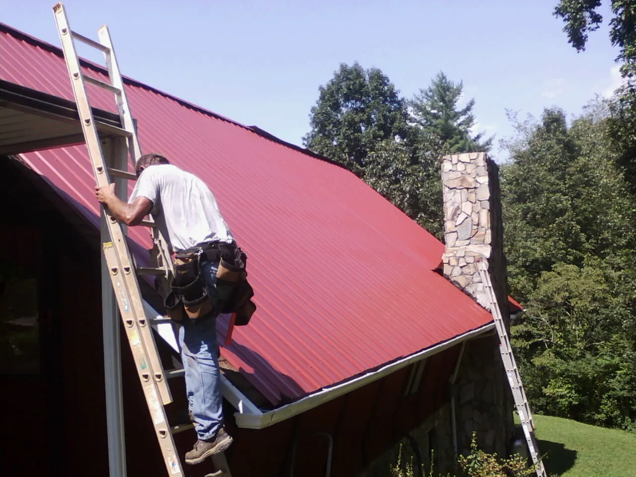 Roofer on a ladder beside a red metal roof and stone chimney — Soffit & Fascia Repair by Roofing Brothers in Romayor